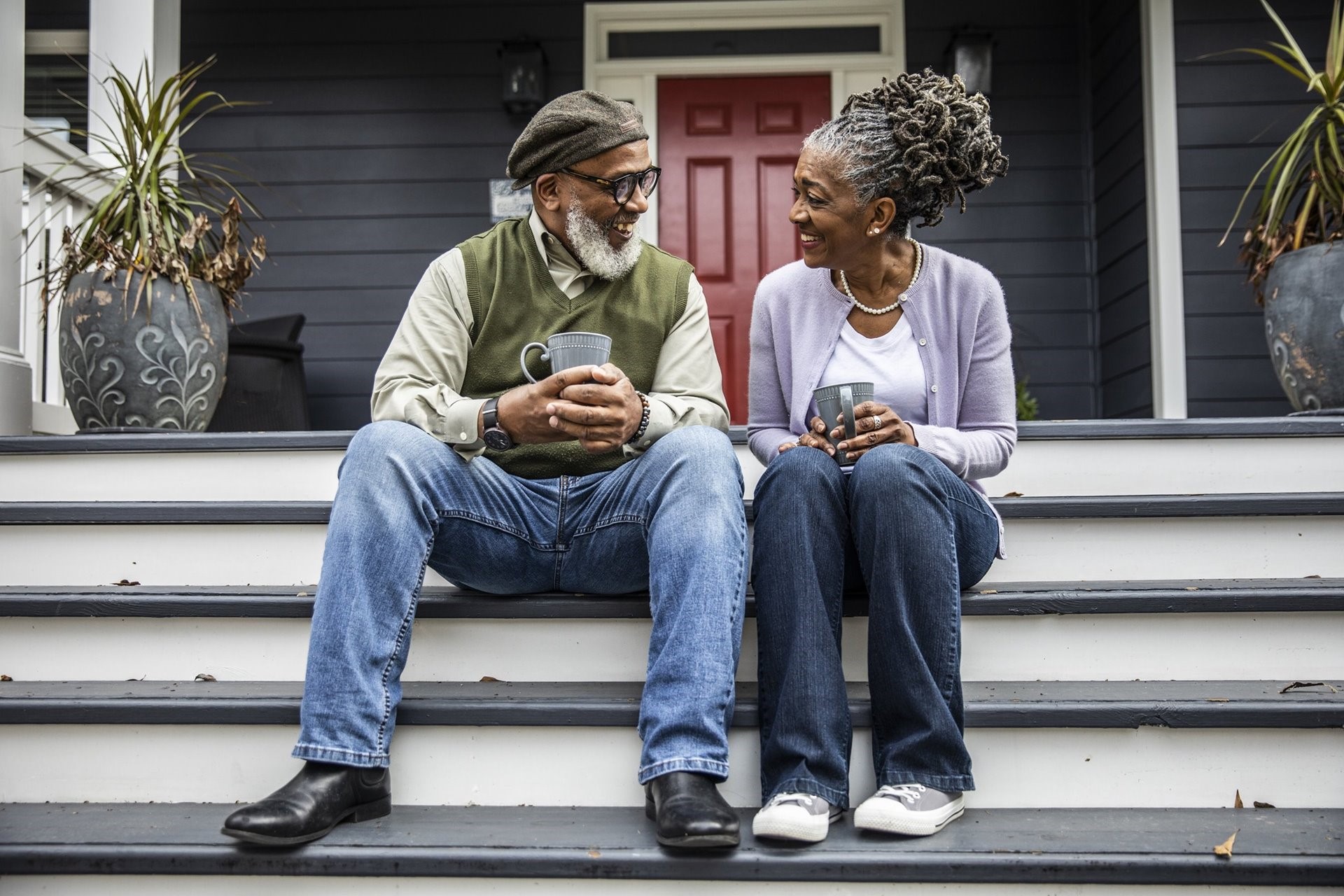 Two elderly couple sitting outside front step smiling and drinking coffee