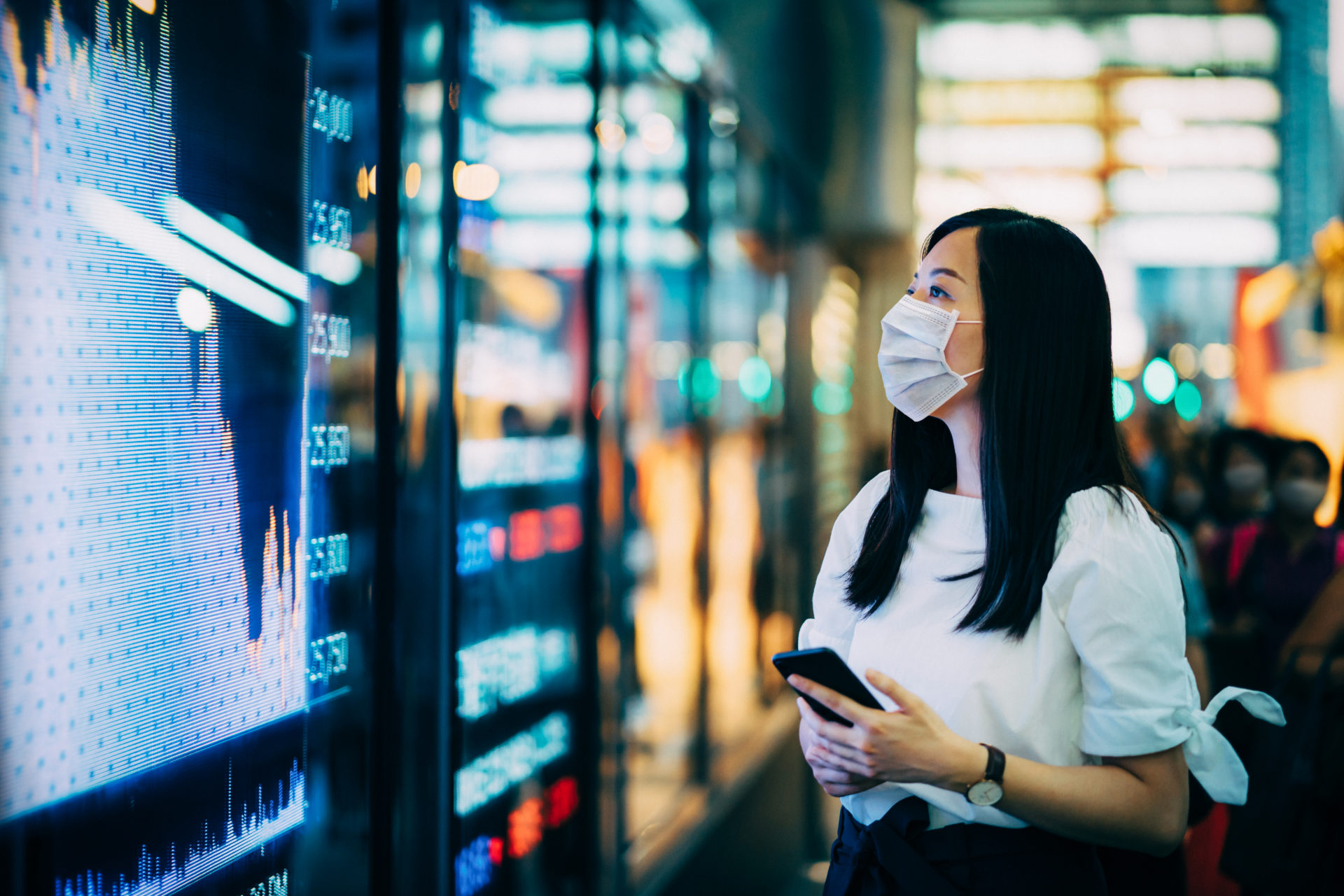 Economic and financial impact during the Covid-19 health crisis deepens. Businesswoman with protective face mask checking financial trading data on smartphone by the stock exchange market display screen board in downtown financial district showing st