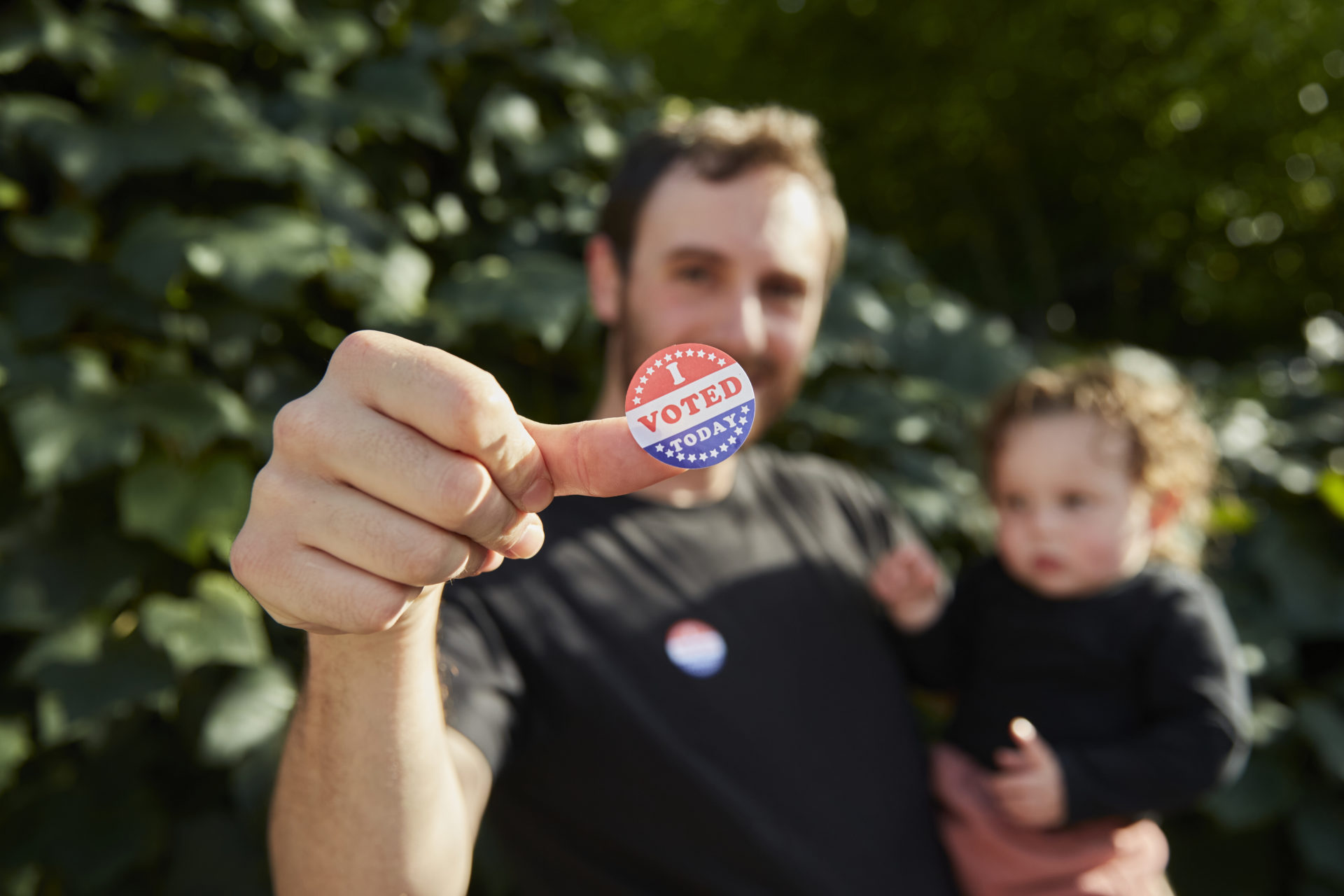 A father standing outside holding his baby, with an "I Voted Sticker" on his thumb