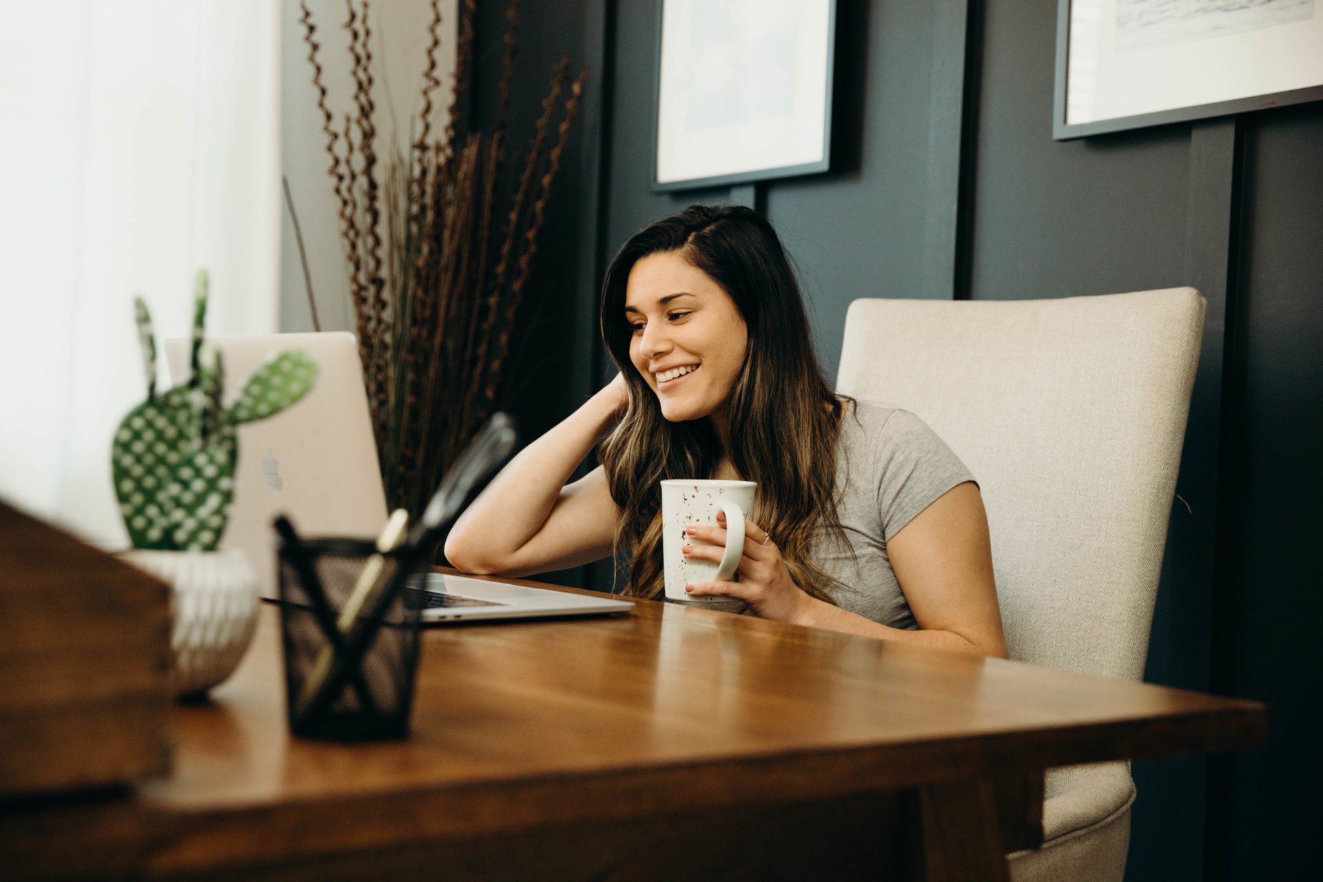 woman with dark hair drinking coffee and smiling at her laptop