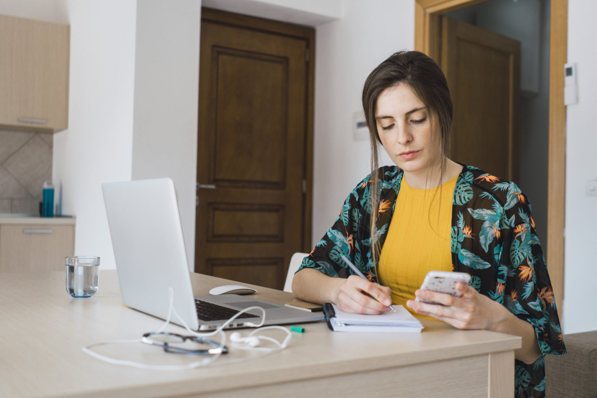 Young woman sitting at table at home using cell phone and laptop taking notes