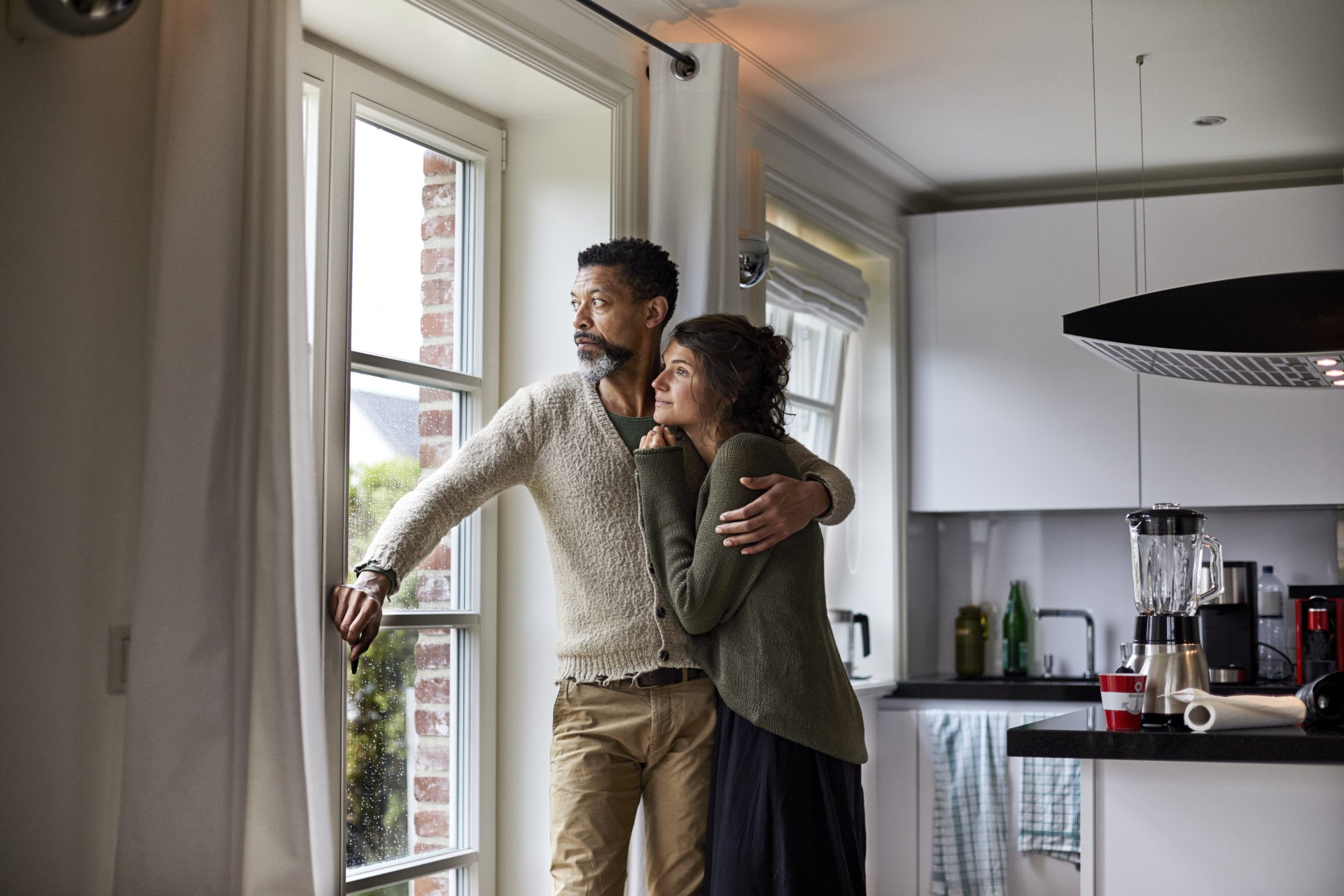 Pensive man embracing young woman looking out of window in kitchen