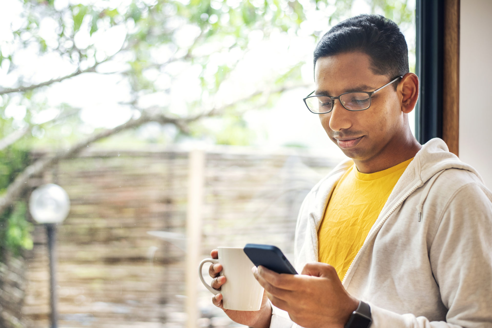 Smiling man drinking coffee and checking his phone by a window at home