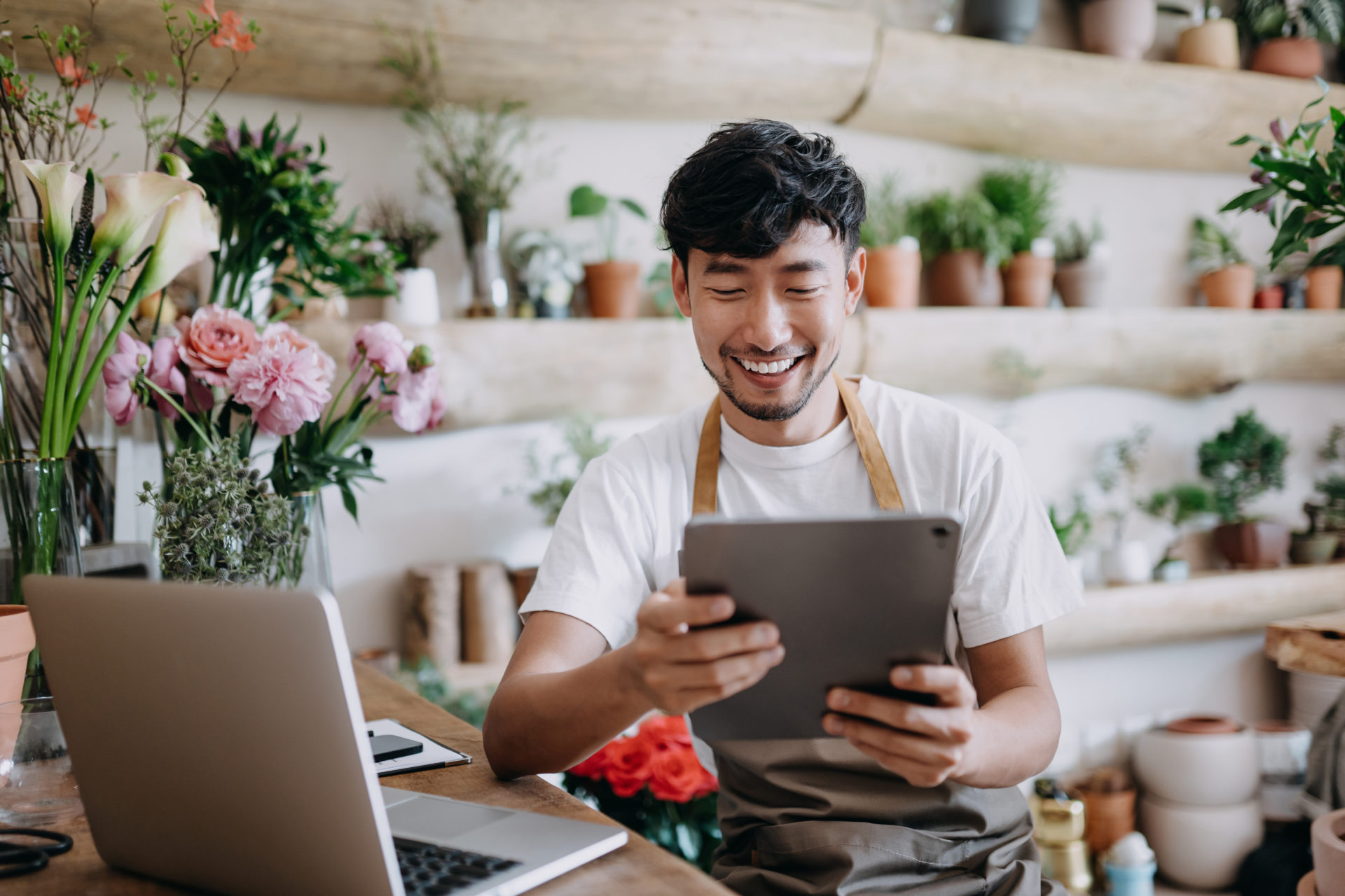 Asian male florist, owner of small business flower shop, using digital tablet while working on laptop against flowers and plants. Checking stocks, taking customer orders, selling products online. Daily routine of running a small business with technol