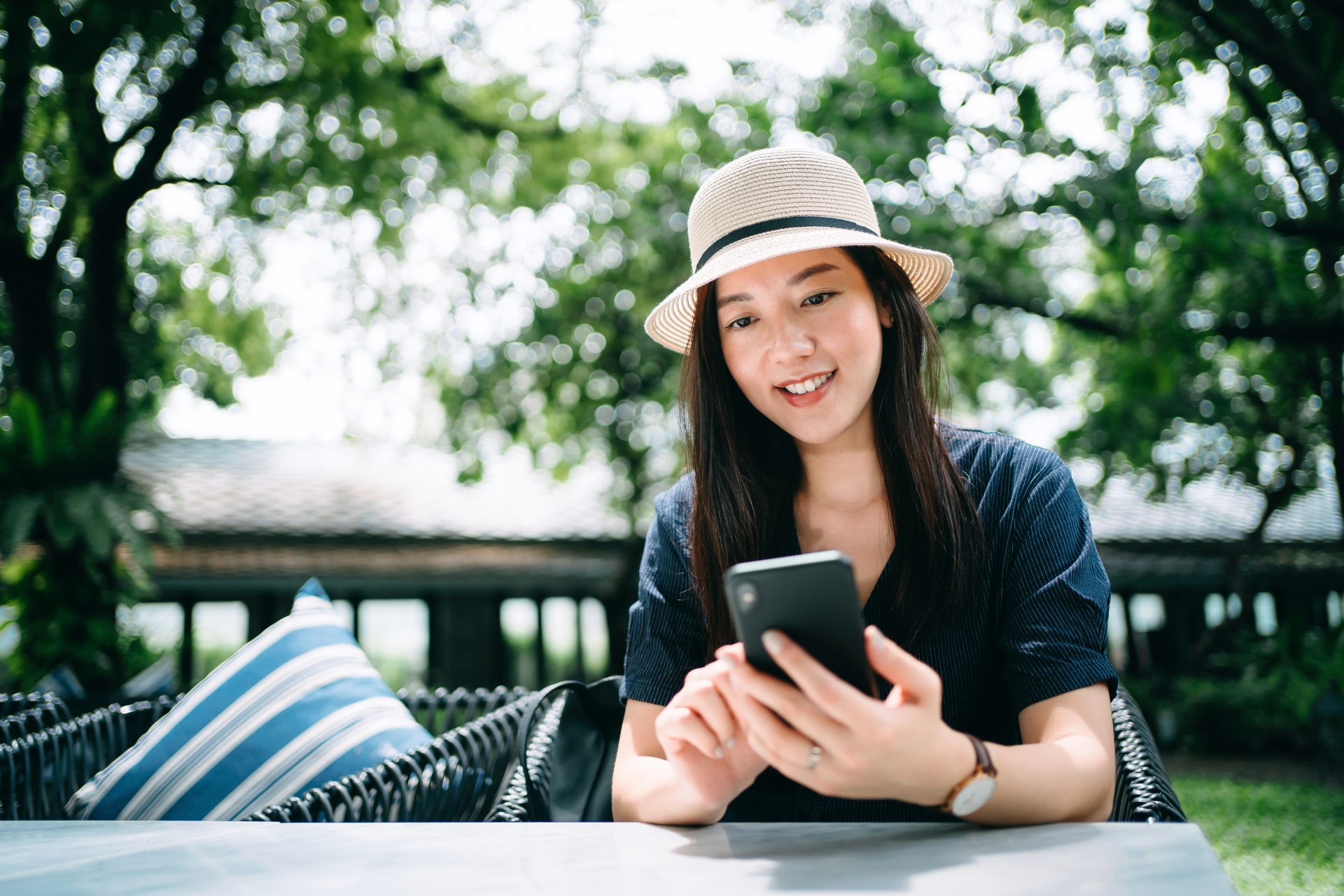 Beautiful young Asian female traveller with a straw hat surfing on the net while relaxing on lounge chair in an outdoor garden at luxury tropical resort