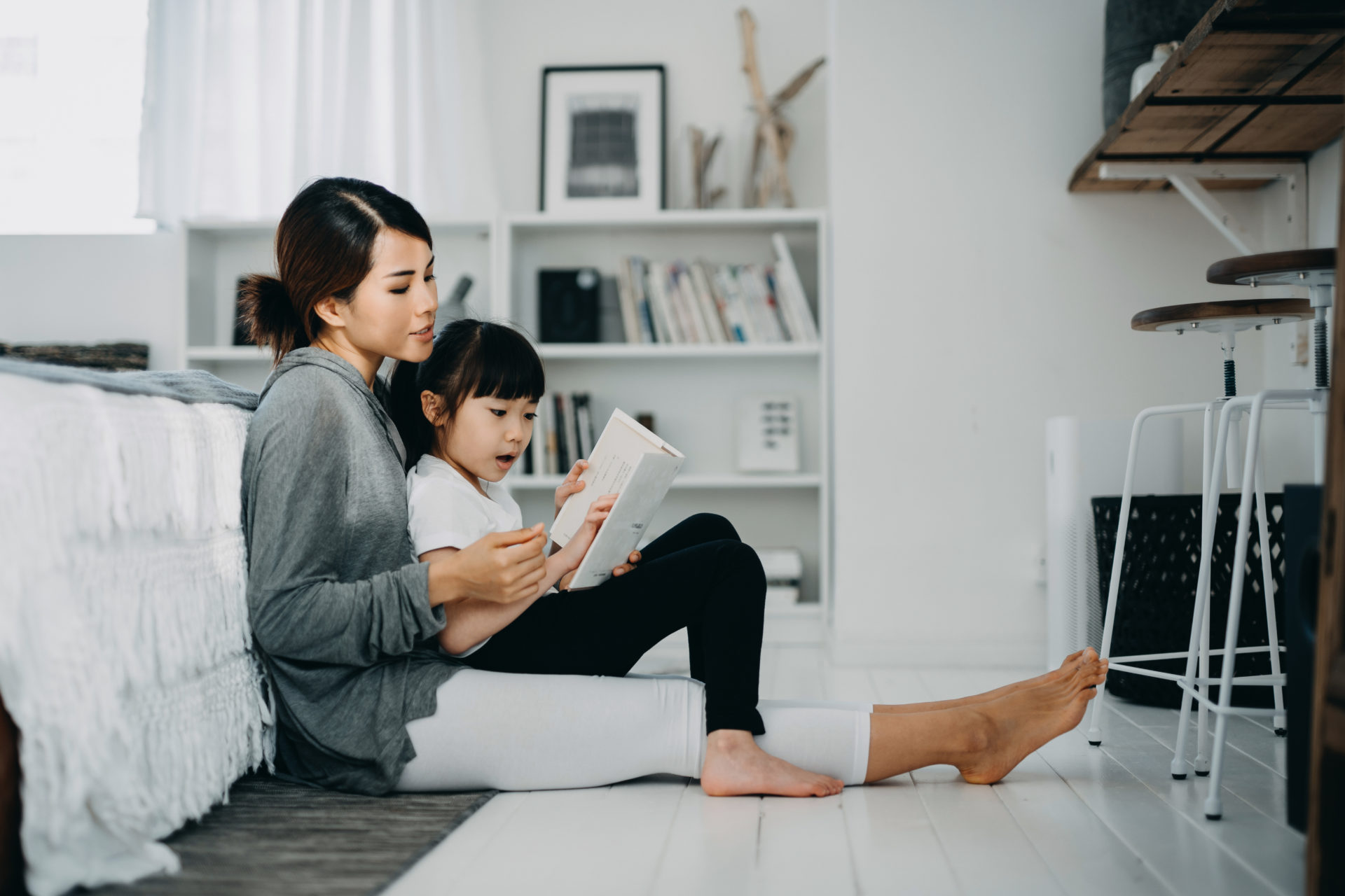 Young Asian mother sitting on the floor in the bedroom reading book to little daughter, enjoying family bonding time together at home