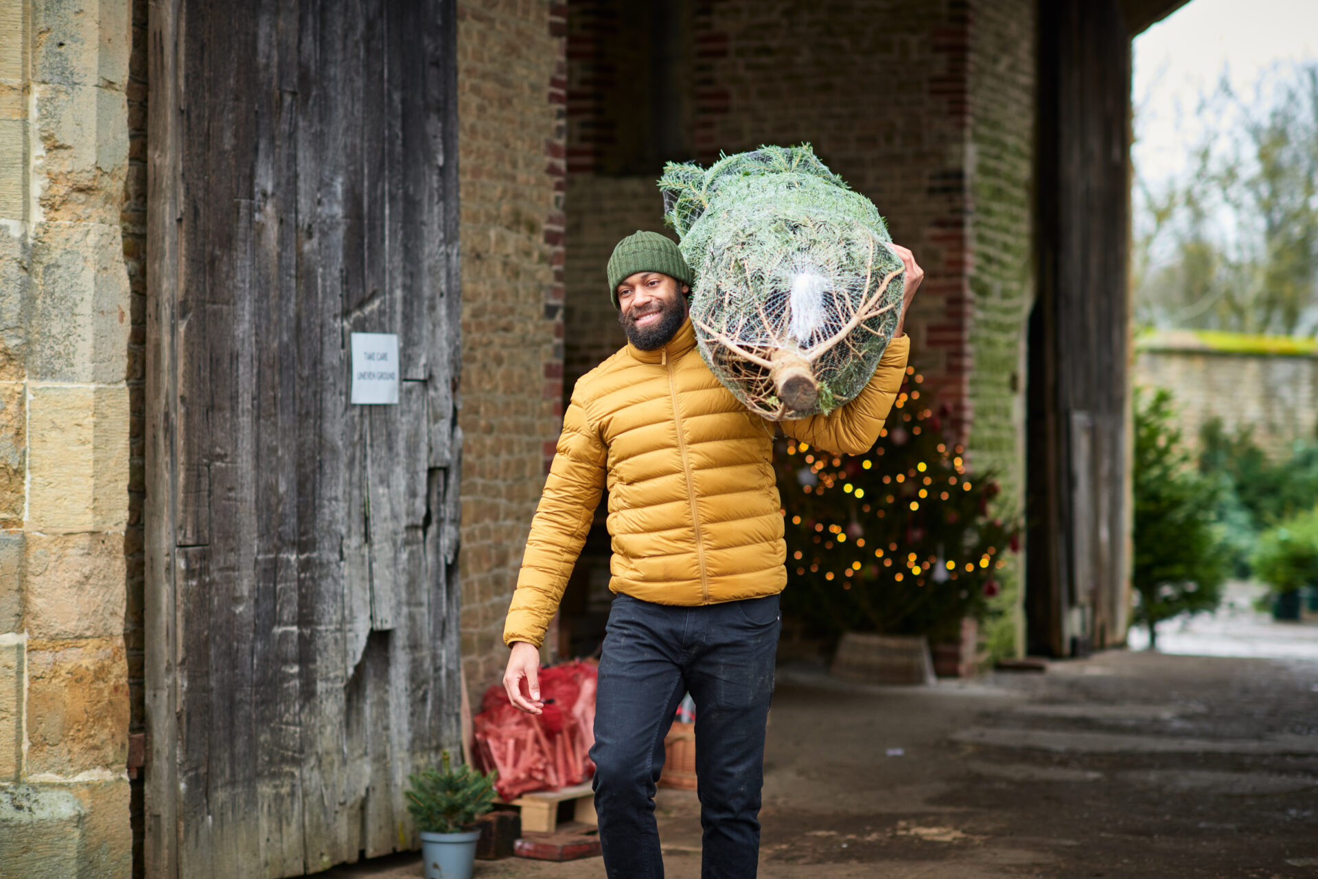 Smiling mid adult black man wearing puffer jacket carrying large wrapped Christmas tree on shoulder