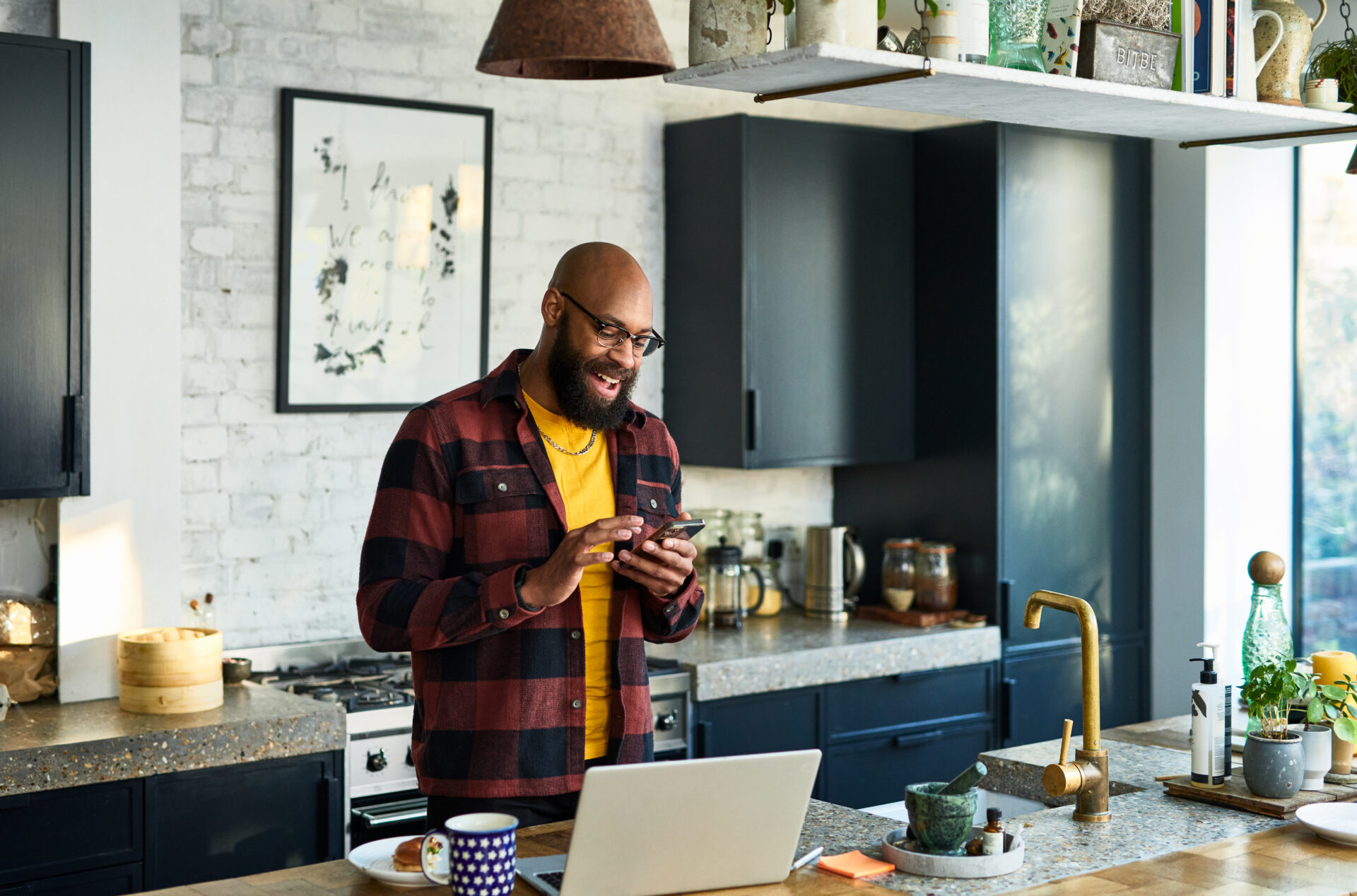 Cheerful black man in his 30s using cell phone in kitchen with laptop on work surface