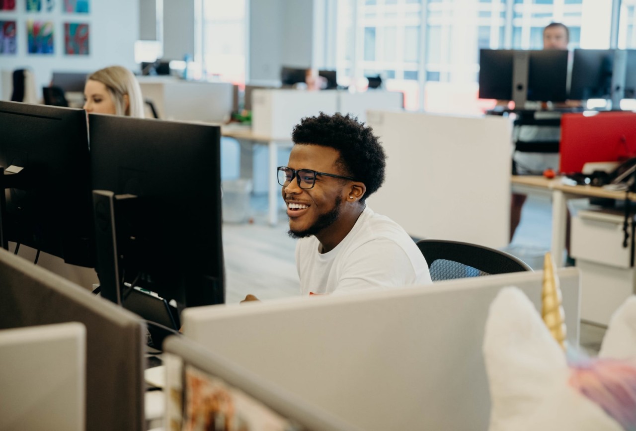 Man with glasses seated in a low-walled cubicle farm looking at two screens and laughing