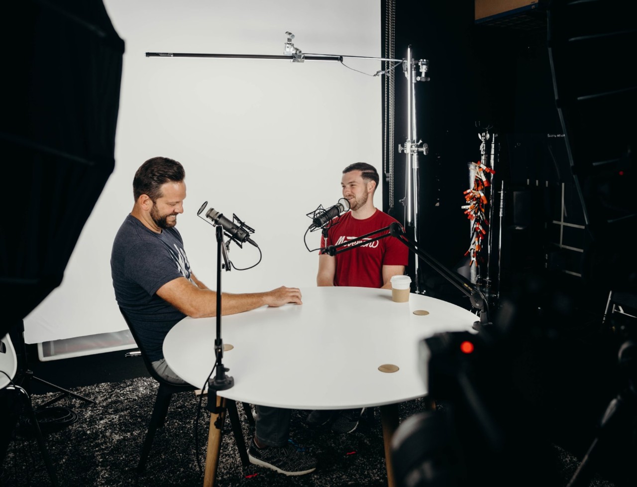 Two men seated at a small table facing each other laughing; working on a podcast