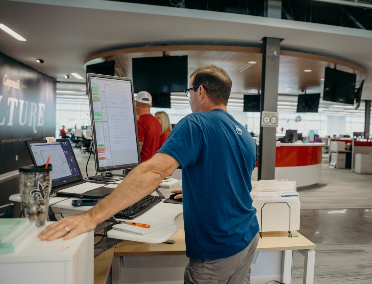 Man, facing away from camera, standing at a corner desk and reading information from a computer monitor in a vertical position